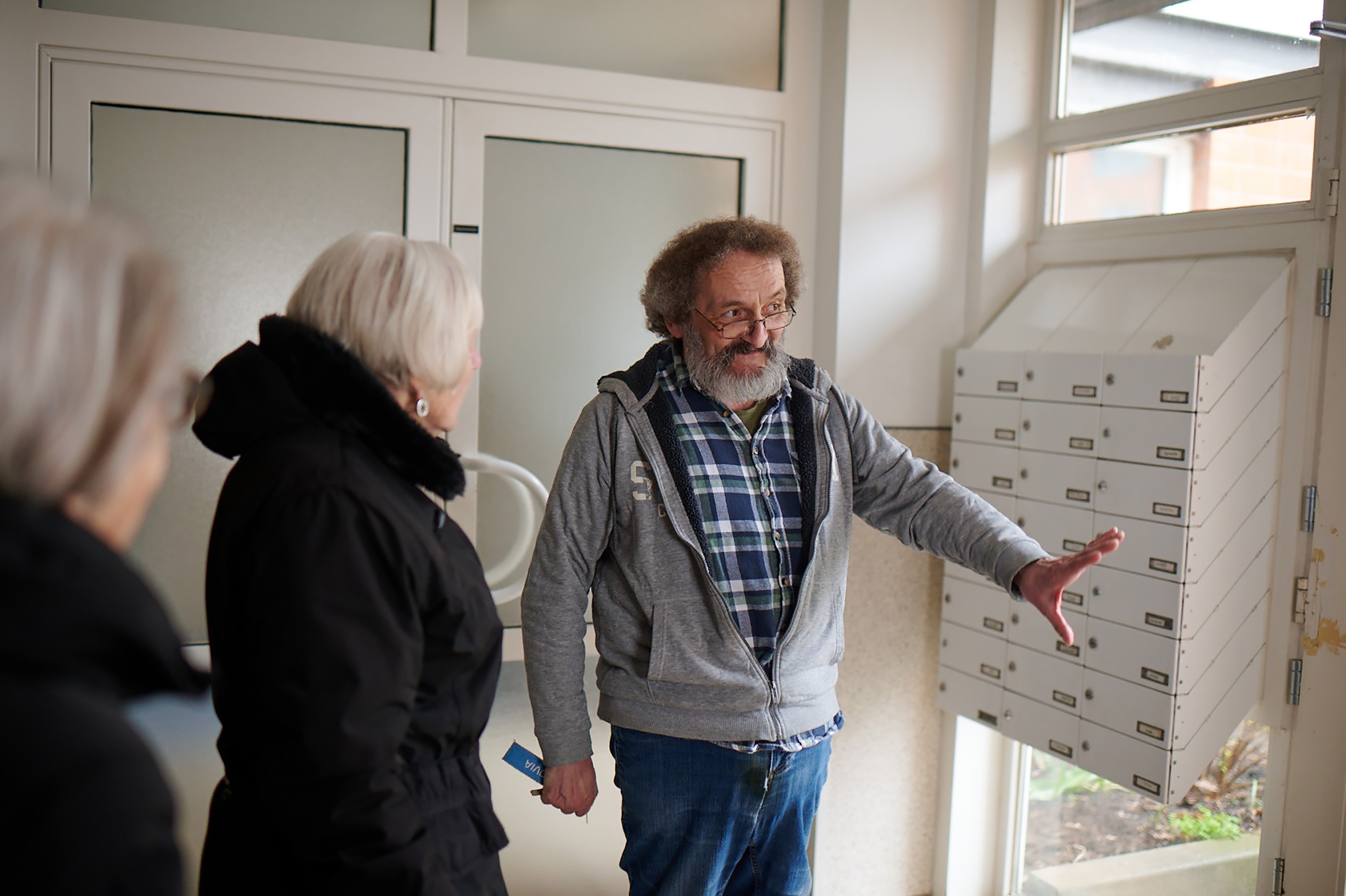 Älterer Mann zeigt Frauen etwas neben Briefkästen im Hausflur.