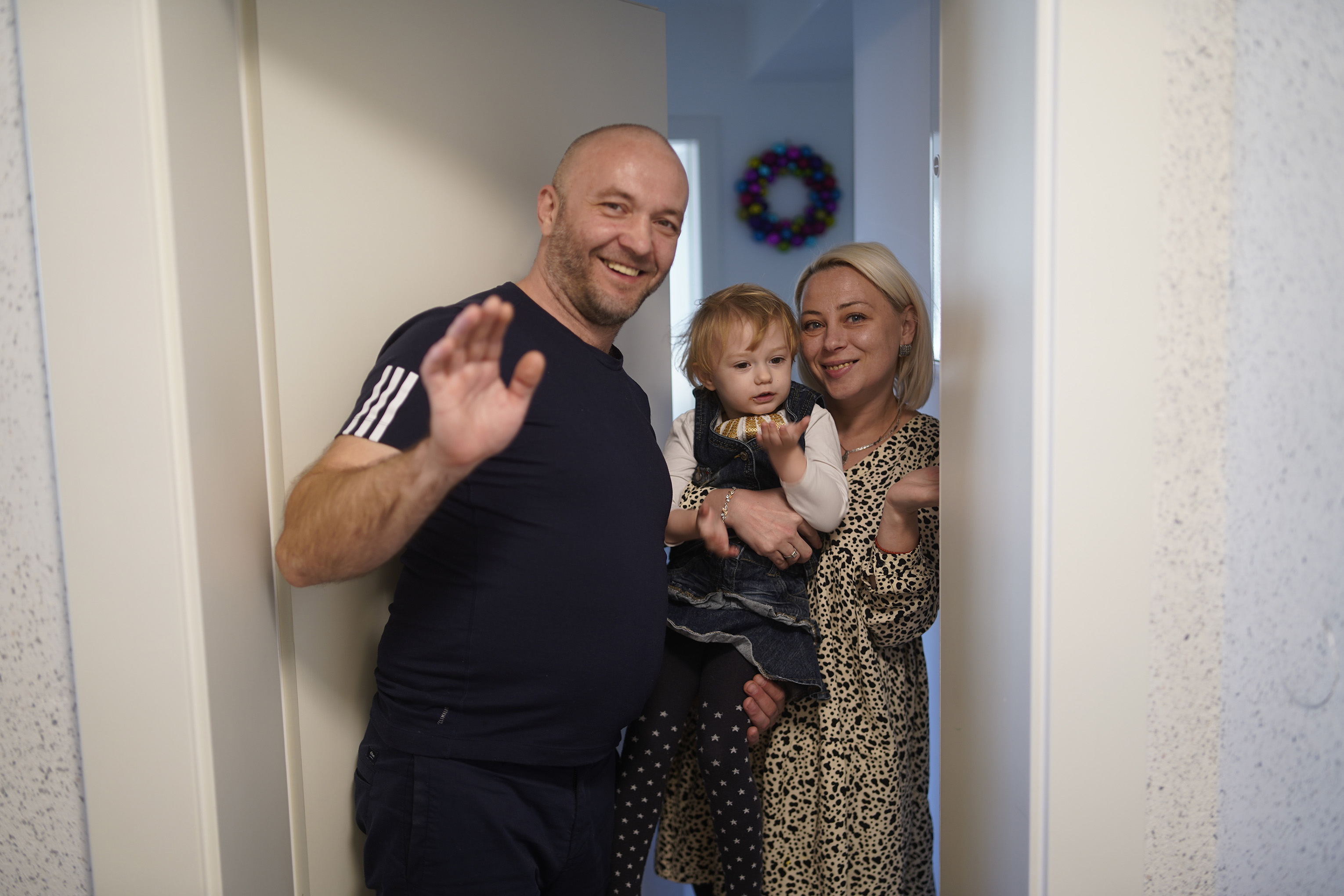 Mann winkt, Frau hält Kind im Türrahmen, Innenraum mit Kranz.