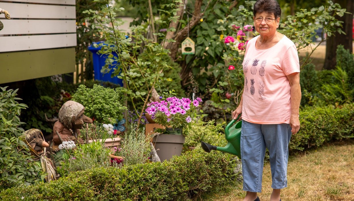 Eine ältere Frau steht lächelnd mit einer grünen Gießkanne in ihrem üppig bepflanzten, mit Dekofiguren geschmückten Garten.