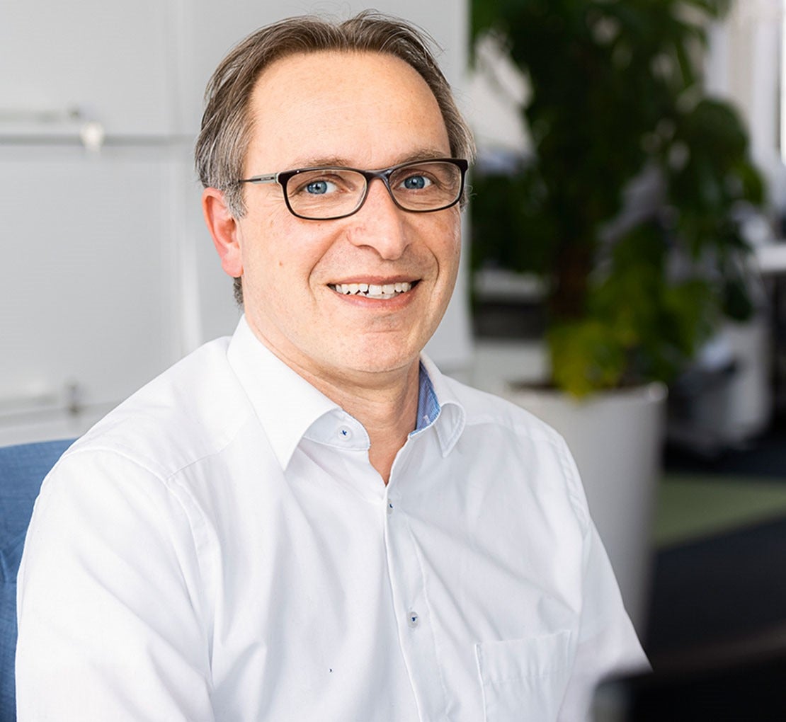 Mann mit Brille lächelt im Büro mit Pflanzen im Hintergrund.