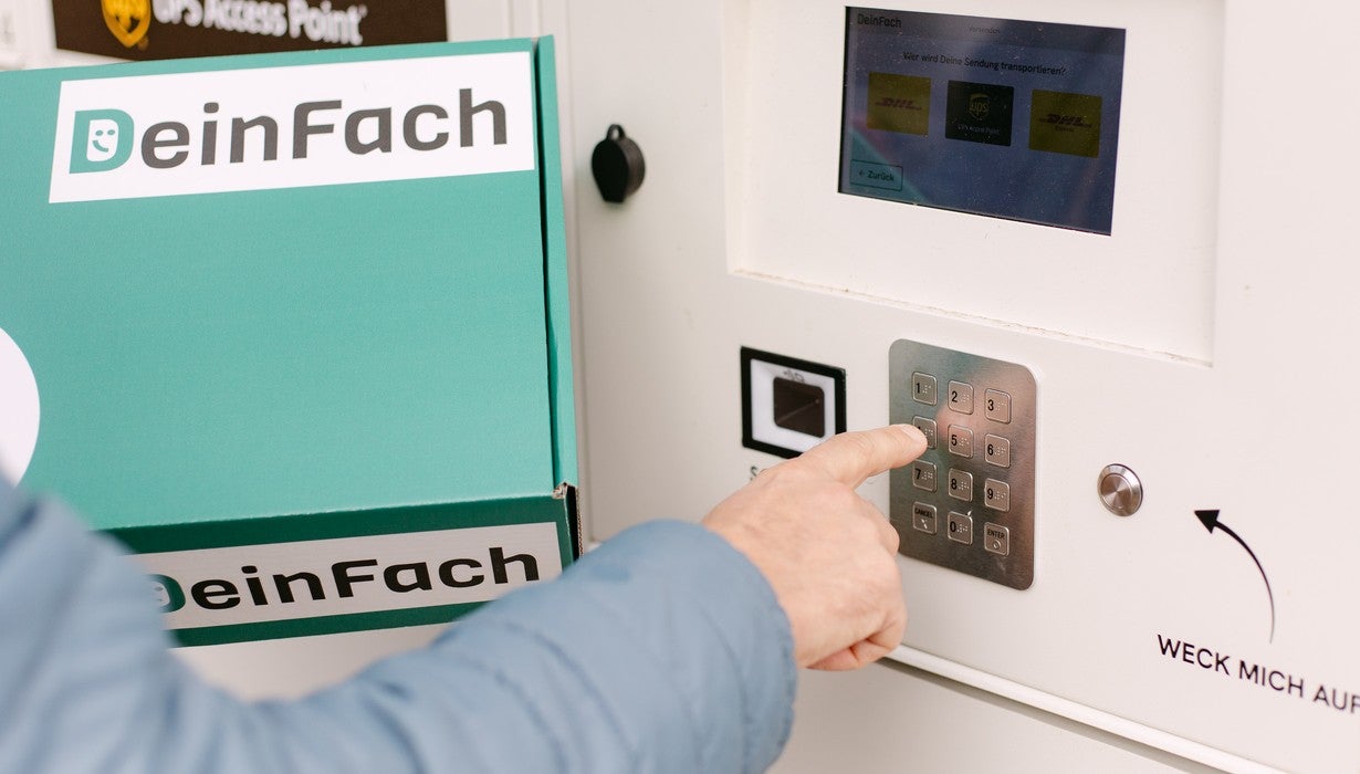 Nahaufnahme einer Hand, die einen Code auf dem Tastenfeld einer „DeinFach“-Paketstation eingibt, während in der anderen Hand ein grünes Paket gehalten wird.