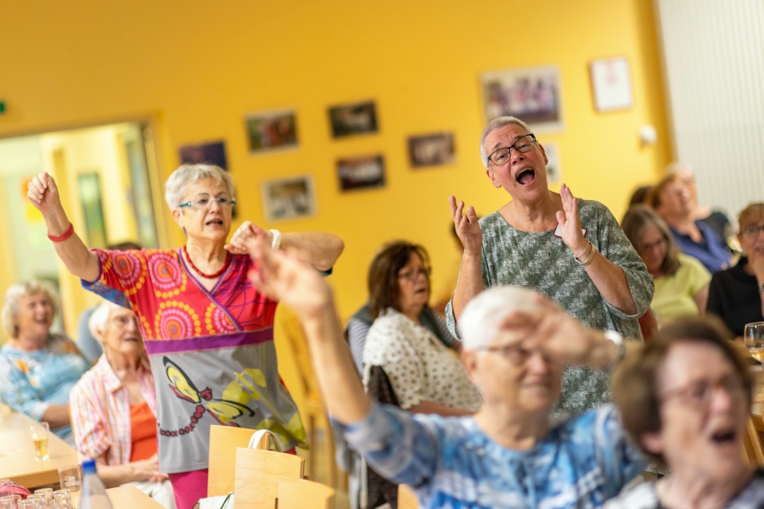 Viele Senioren singen zusammen, zwei Personen im Fokus, drumherum Weitere, alle klatschen oder bewegen die Arme.