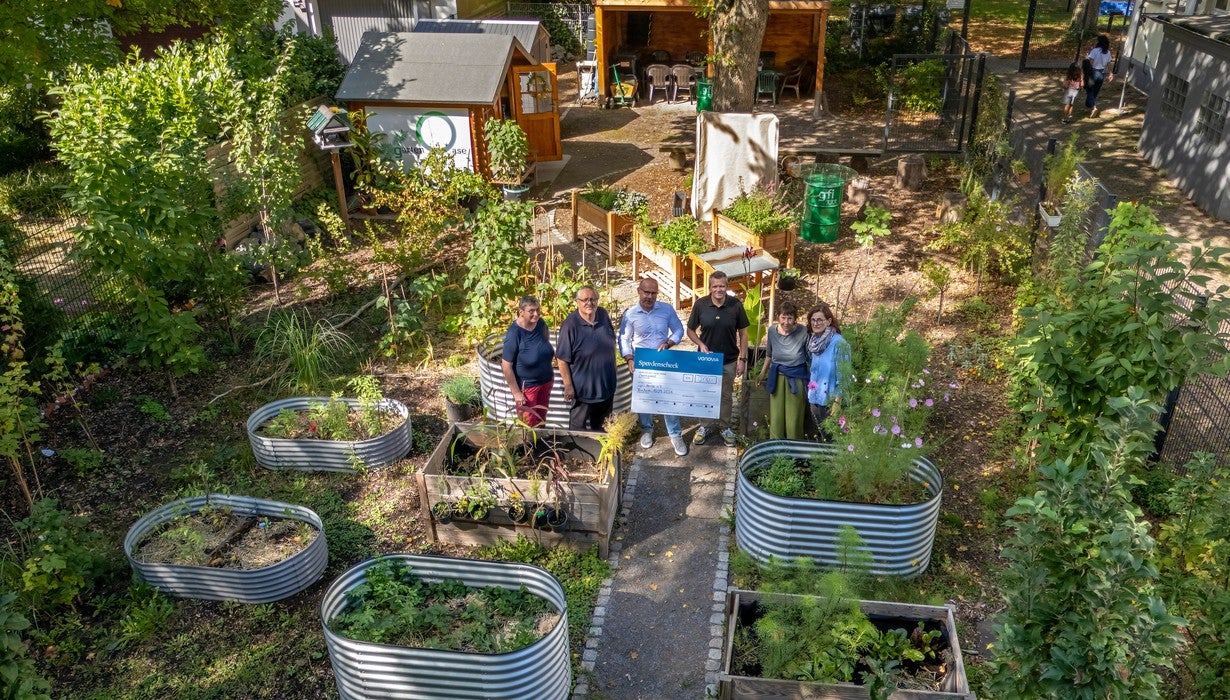 Eine Gruppe von Menschen präsentiert gemeinsam einen Vonovia-Spendenscheck inmitten eines üppig bepflanzten Gemeinschaftsgartens mit Hochbeeten und einem kleinen Gartenhaus aus der Vogelperspektive.