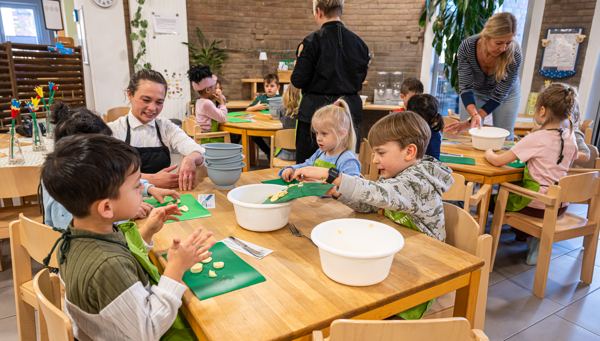 Kinder sitzen gemeinsam mit Betreuerinnen an Holztischen in einem Raum und bereiten Essen zu, indem sie Obststücke von grünen Schneidbrettern in weiße Schüsseln füllen.