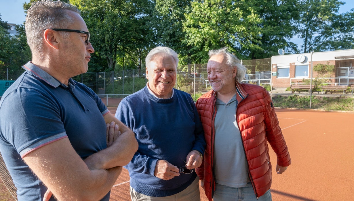 Drei Männer unterhalten sich gut gelaunt am Rand eines Tennisplatzes, wobei zwei von ihnen lächeln und einer eine rote Steppjacke trägt.