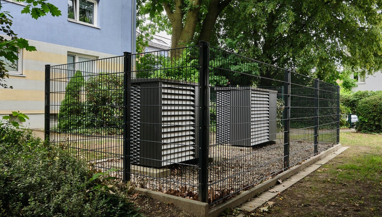 Zwei moderne Wärmepumpen stehen eingezäunt in einem mit Metallzaun umgrenzten Außenbereich neben einem Wohngebäude im Grünen.