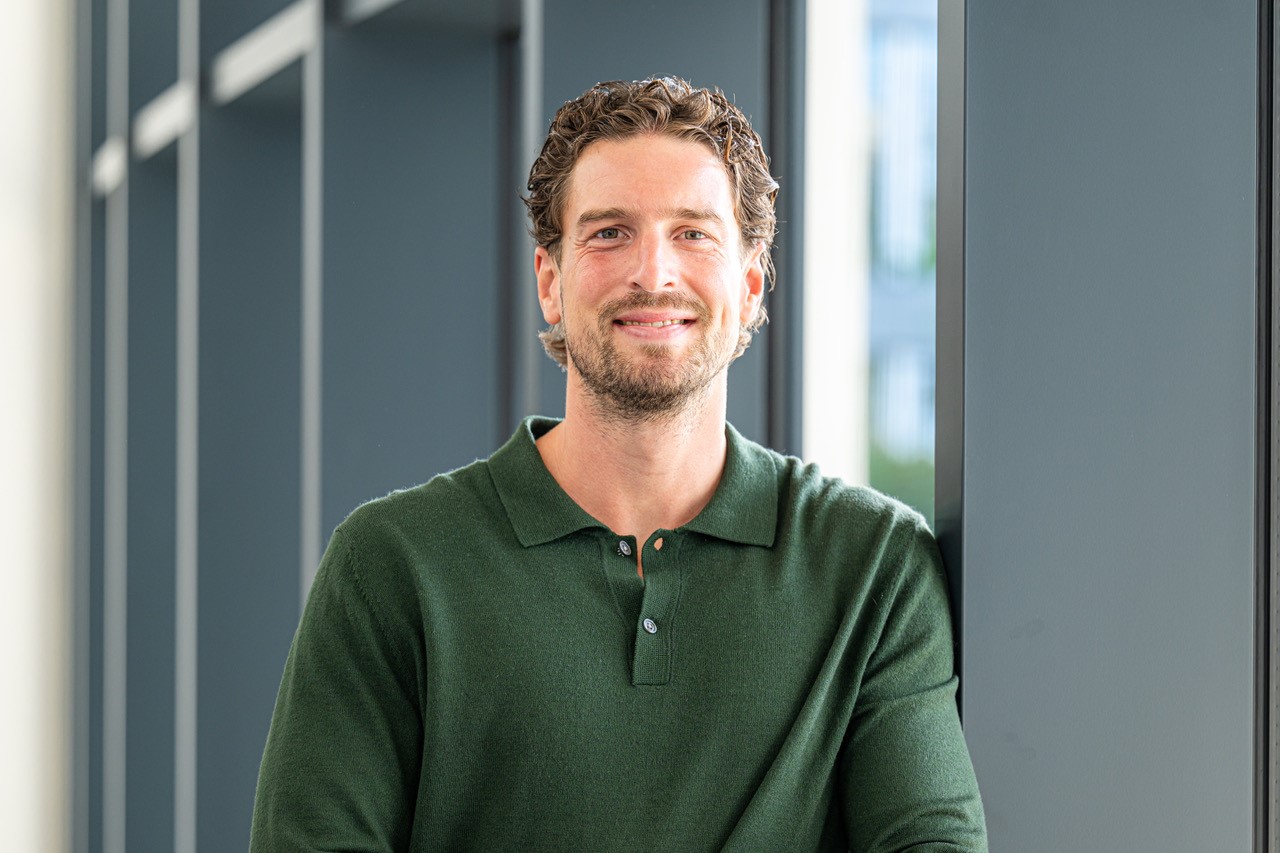 Portrait Konstantin Fassbender. Lächelnder Mann mit lockigem, braunem Haar und Bart. Er trägt ein dunkelgrünes Polo-Shirt und lehnt an einen grauen Balken. Portrait Konstantin Fassbender. Lächelnder Mann mit lockigem, braunem Haar und Bart. Er trägt ein dunkelgrünes Polo-Shirt und lehnt an einen grauen Balken.