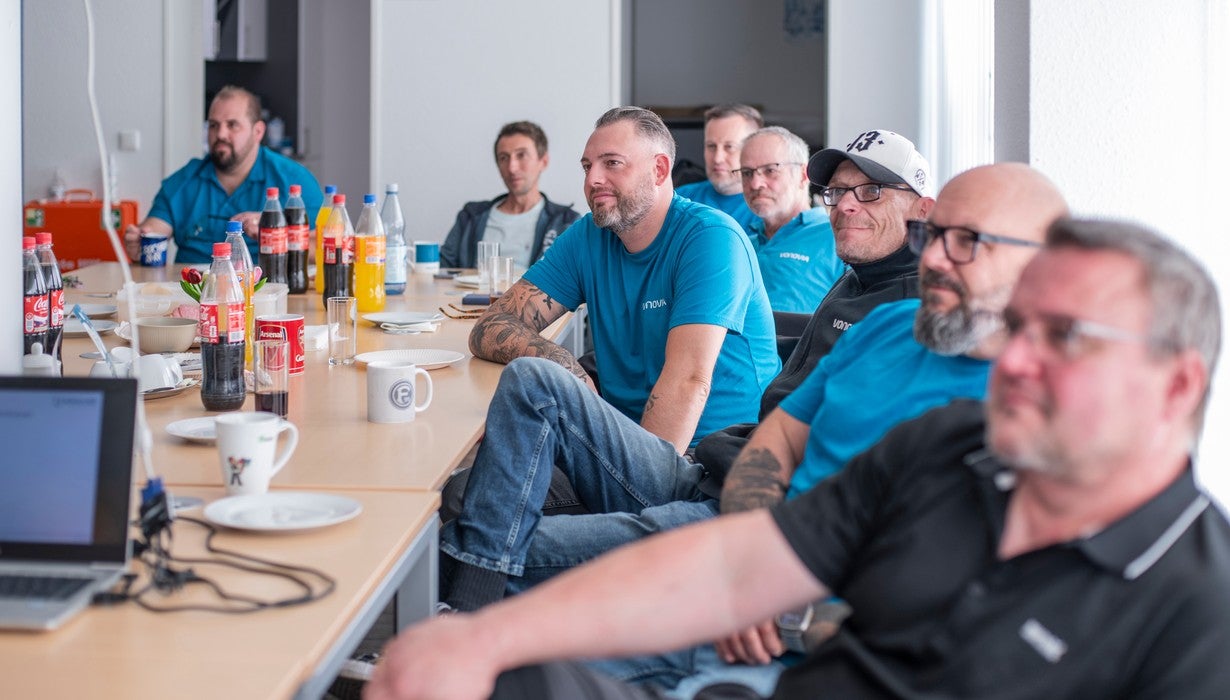 Eine Gruppe von Männern sitzt an einem langen Tisch mit Getränken während einer Besprechung oder Schulung in einem Innenraum und blickt aufmerksam nach vorne.