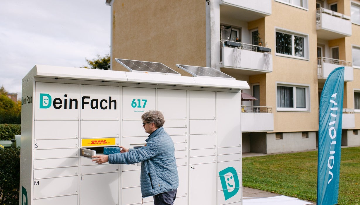 Ein Mann legt ein Paket in ein Fach einer weißen, solarbetriebenen Paketstation namens „DeinFach“ vor einem mehrstöckigen Wohngebäude mit einer blauen Vonovia-Fahne im Hintergrund.
