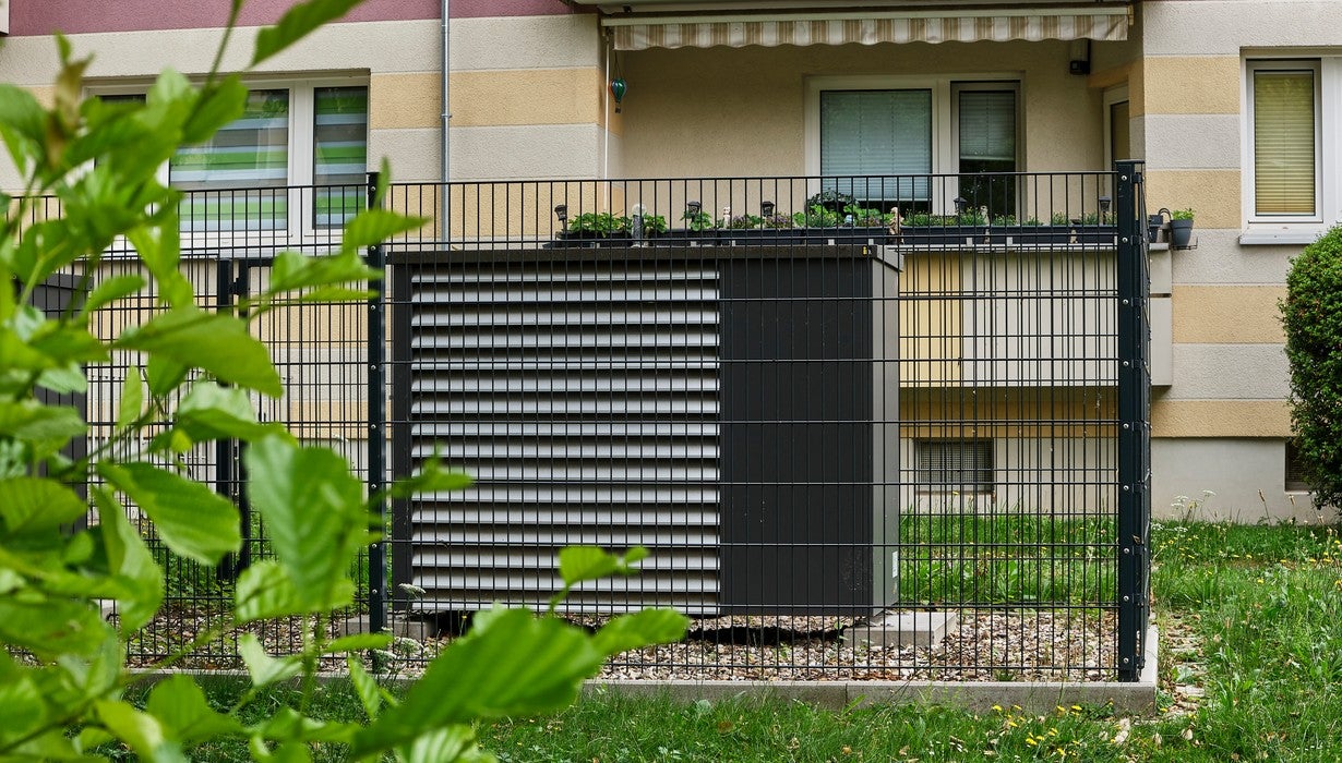 Eine moderne Wärmepumpe steht hinter einem dunklen Metallzaun vor der Fassade eines Wohngebäudes.
