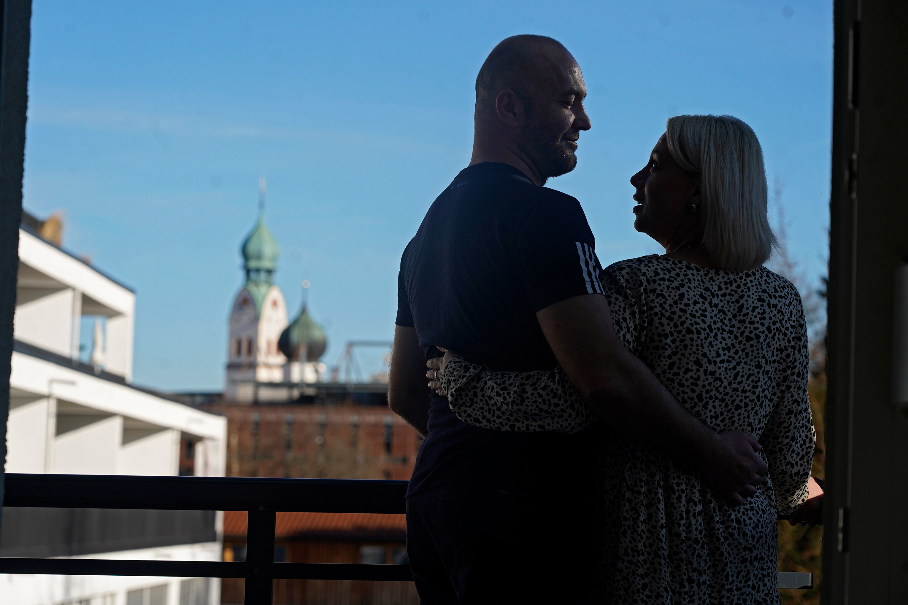 Eine Frau und ein Mann stehen auf einem Balkon, halten sich in den Armen und schauen sich an. Man sieht unscharf die Umrisse der umliegenden Gebäude und eine Kirche. Eine Frau und ein Mann stehen auf einem Balkon, halten sich in den Armen und schauen sich an. Man sieht unscharf die Umrisse der umliegenden Gebäude und eine Kirche.
