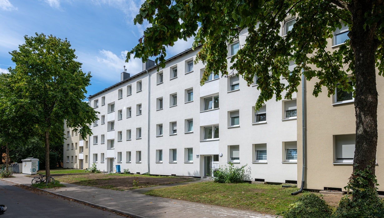 Ein mehrstöckiges, weiß-gelb gestrichenes Wohngebäude an einer ruhigen Straße mit schattenspendenden Bäumen und gepflegten Grünflächen davor.