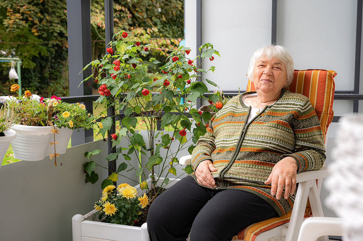 Seniorin sitzt auf einem gemütlichen Stuhl auf einem Balkon. Daneben steht ein Pflanzkasten mit Blumen und am Geländer hängen weitere Pflanzkübel.