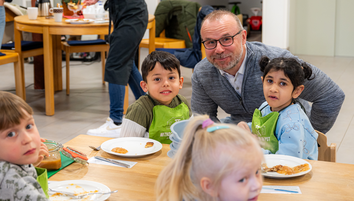 Ein lächelnder Mann mit Brille lehnt sich zu zwei Kindern in grünen Kochschürzen, die gemeinsam an einem Holztisch sitzen und essen.