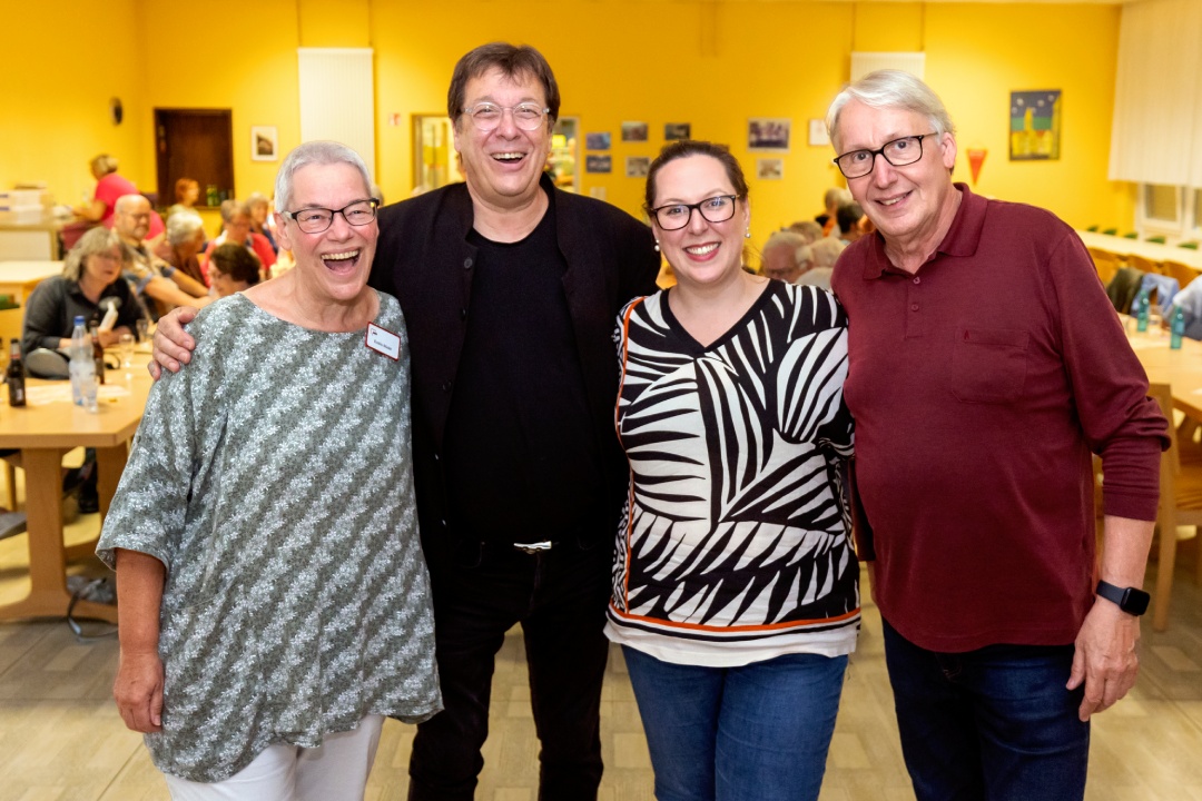 4 Seniorinnen und Senioren posieren für ein Foto, im Hintergrund Tische an denen andere Personen sich unterhalten.
