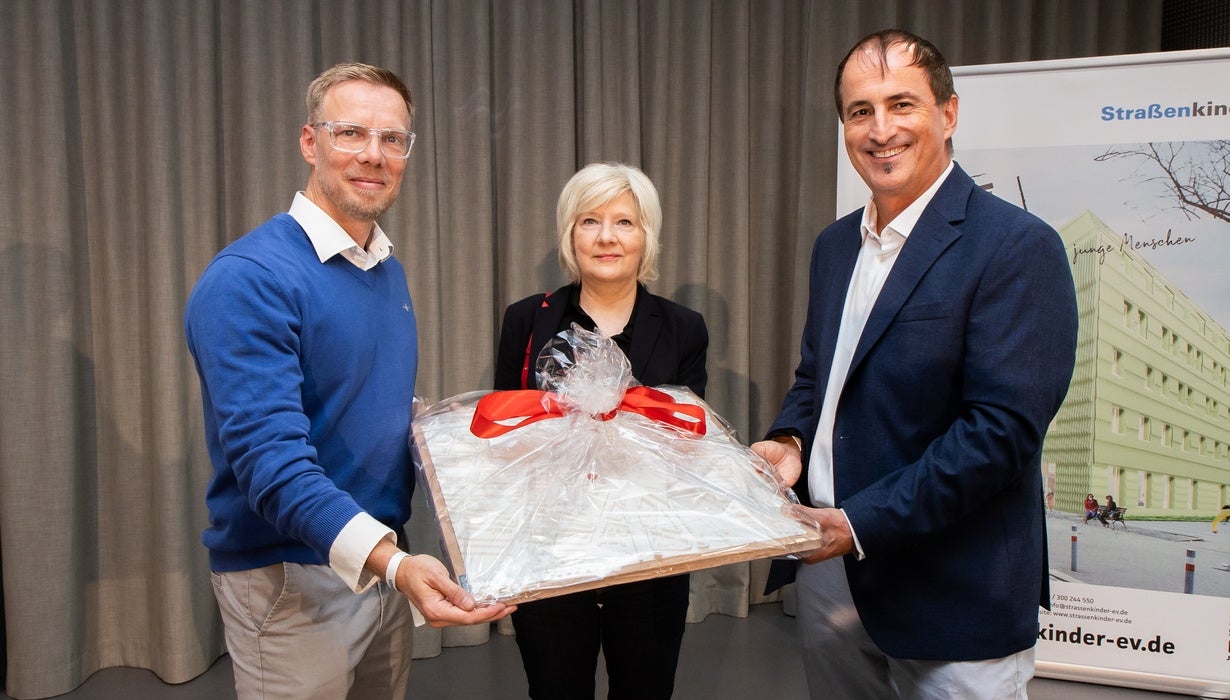 Drei Personen übergeben vor einem Vorhang und dem Banner von „Straßenkinder e.V.“ gemeinsam ein verpacktes, mit einer roten Schleife verziertes Geschenk.