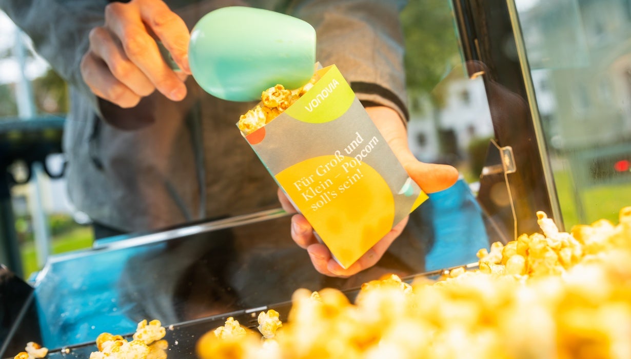 Nahaufnahme einer Hand, die mit einer hellgrünen Schaufel frisches Popcorn in eine bunte Tüte mit Vonovia-Logo und der Aufschrift „Für Groß und Klein – Popcorn soll's sein!“ füllt.