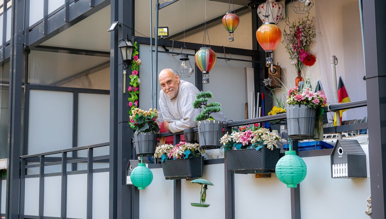 Ein älterer Mann steht auf seinem reich dekorierten Balkon, der mit zahlreichen Blumenkästen, bunten Lampions, Hängeleuchten und einem kleinen Vogelhäuschen geschmückt ist.