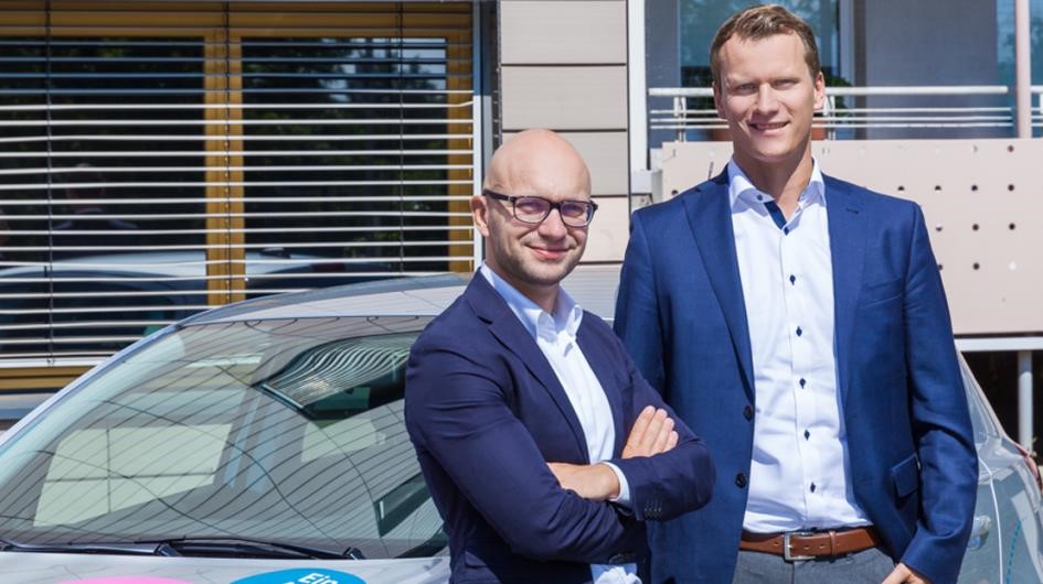 Zwei Männer posieren vor einem Gebäude neben einem Auto.