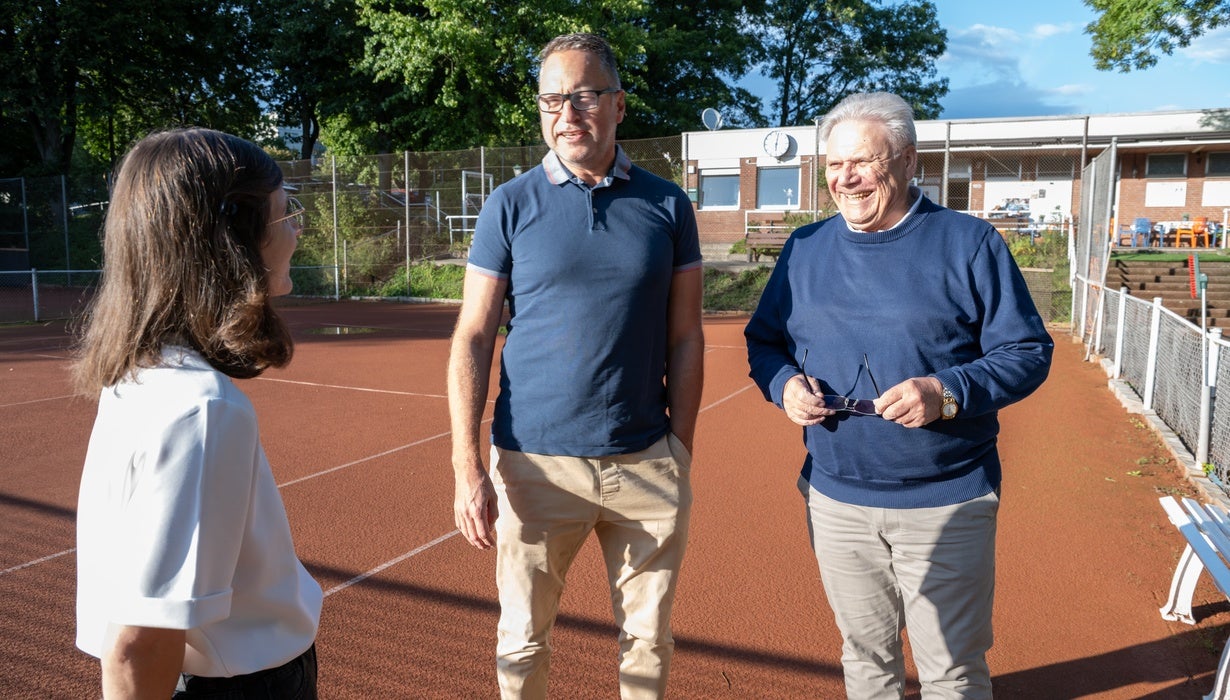 Drei Personen stehen im Gespräch auf einem roten Tennisplatz im Freien, während rechts ein älterer Mann mit Brille in der Hand her herzlich lacht.