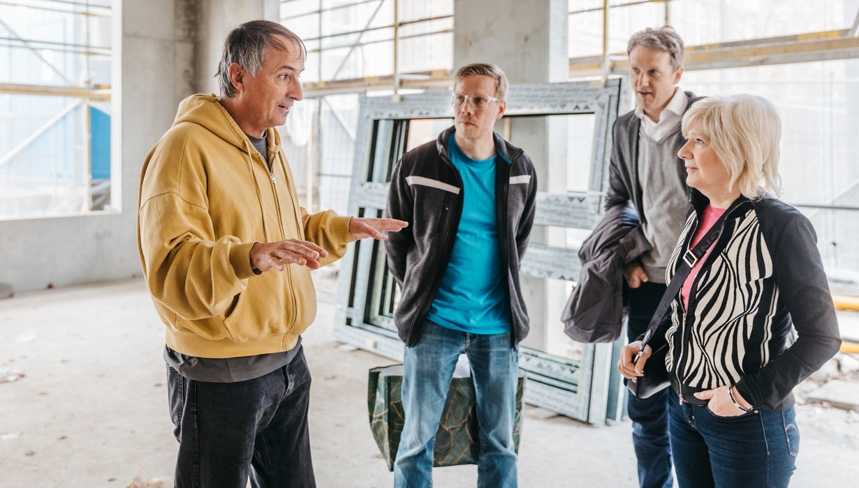 Ein Mann in einer gelben Kapuzenjacke Gestikuliert auf einer Baustelle im Rohbau und erklärt drei aufmerksam zuhörenden Personen das Bauprojekt.