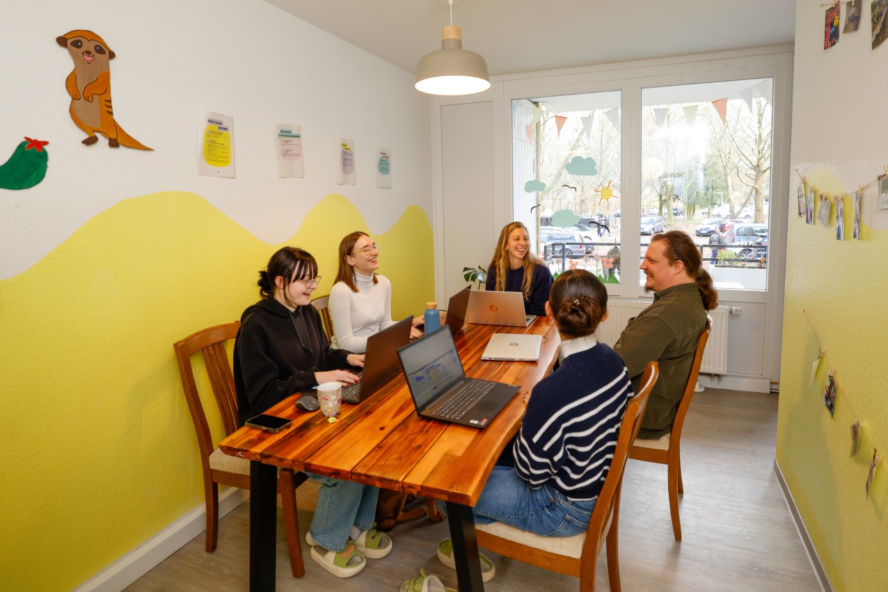 Gruppe von Personen sitzt an einem Tisch in einem hellen Raum mit Wandbemalung & Bildern. Auf dem Tisch sind mehrere Laptops, die Personen reden untereinander.