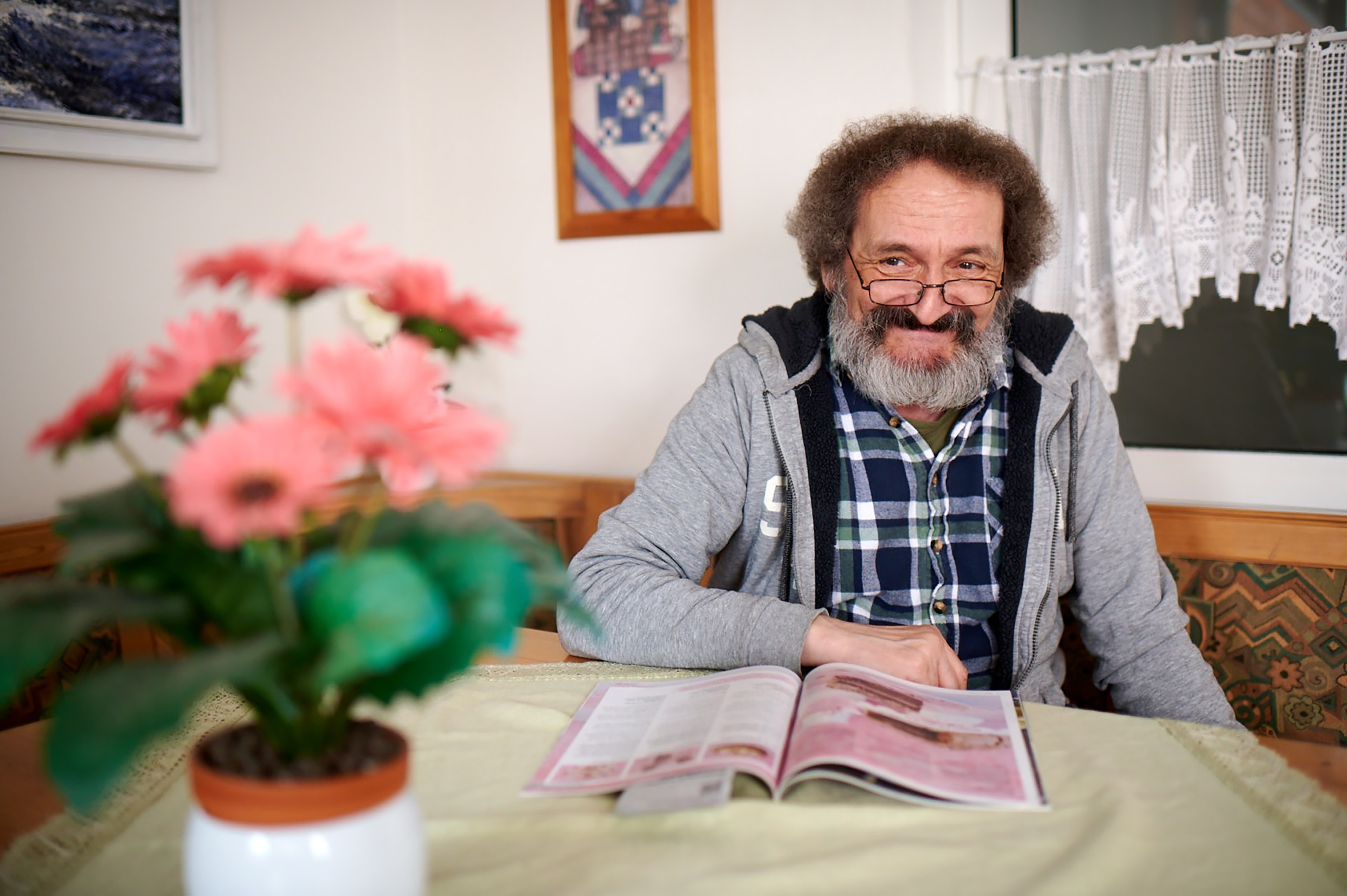 Älterer Mann liest Magazin am Tisch. Blumen im Vordergrund.