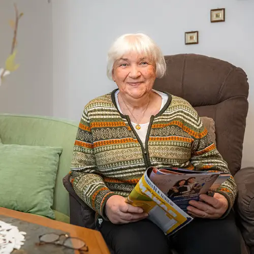 Eine ältere Dame sitzt auf einem Sessel mit einer Zeitschrift in der Hand.