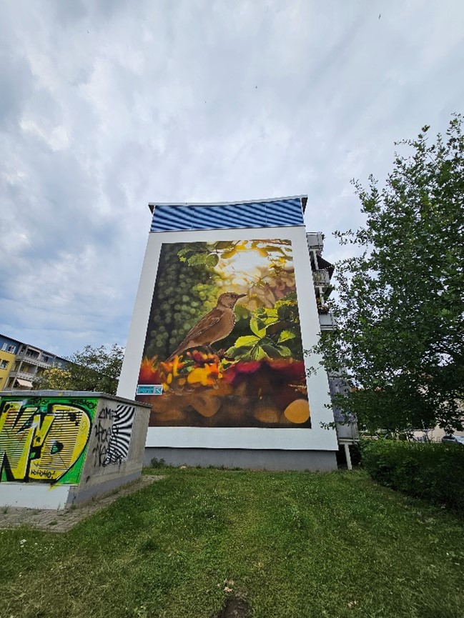 Graffiti von Vogel auf Hauswand, umgeben von Bäumen und Wiese.