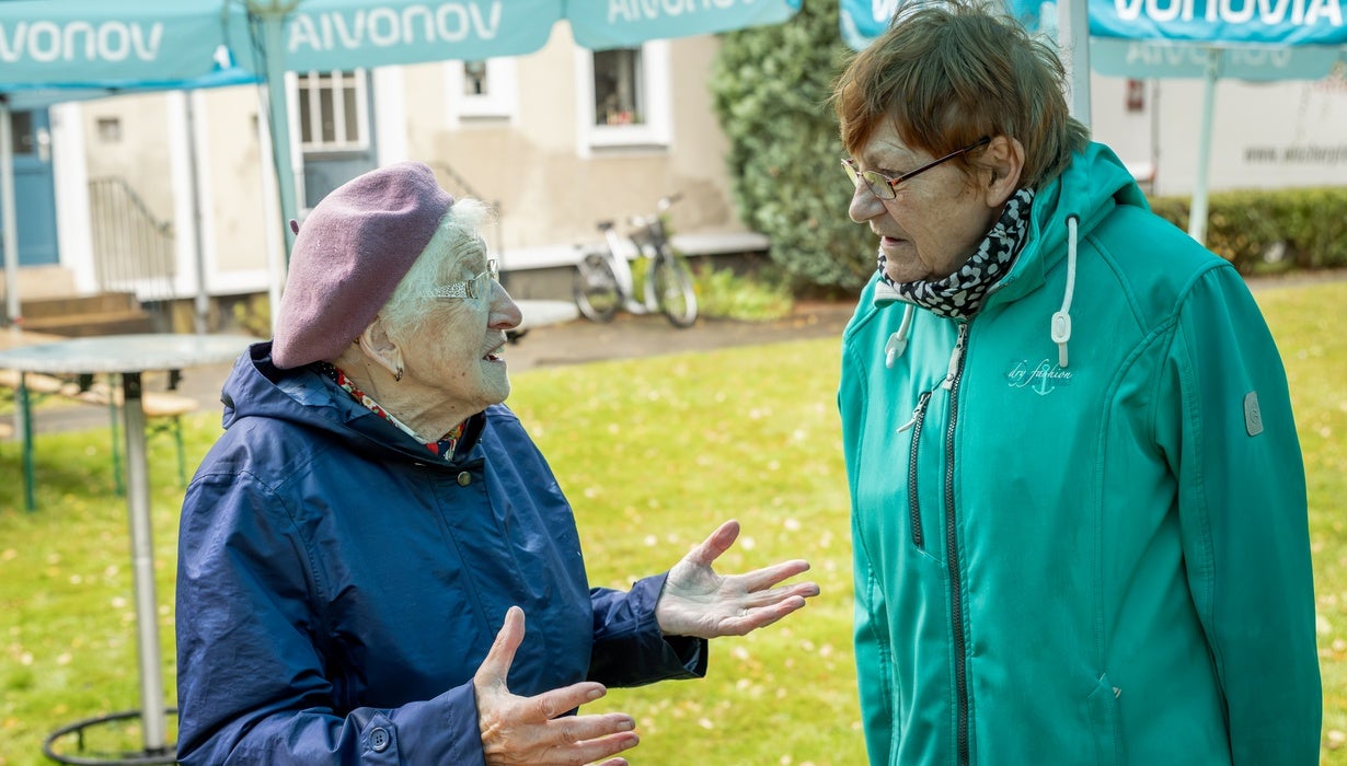 Zwei ältere Frauen unterhalten sich im Freien unter einem Vonovia-Sonnenschirm, während eine von ihnen mit lila Baskenmütze gestikuliert.