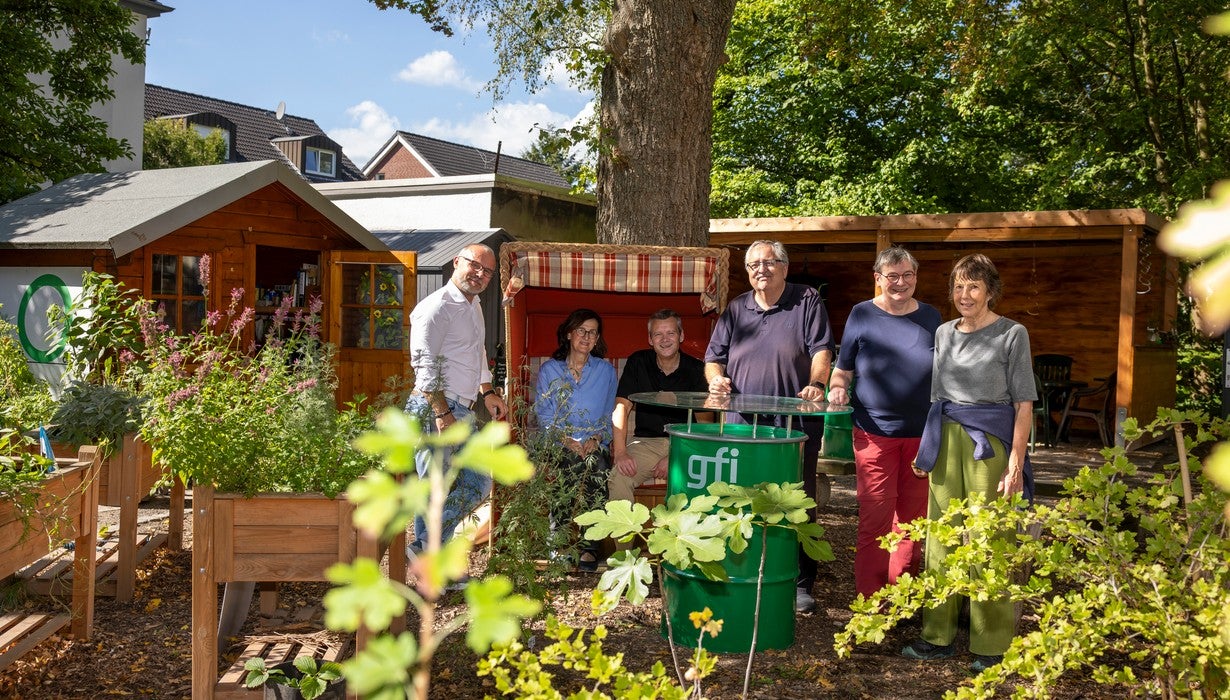 Eine Gruppe von Menschen posiert lächelnd in einem schattigen Gemeinschaftsgarten rund um einen Strandkorb, Stehtisch und ein kleines Gartenhaus im Hintergrund.
