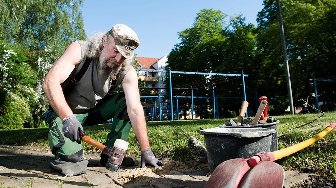 Mann kniet, schlägt Stein, Park mit Spielplatz im Hintergrund.